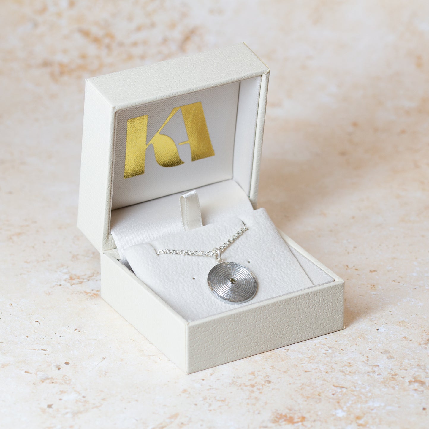 A silver necklace with a circular disc pendant set with a yellow diamond, displayed inside an open cream jewellery box with a gold Kate Archer logo on the lid, set against a light beige textured background.