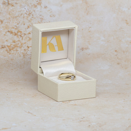 A gold ring with a princess cut diamond set in a shaped-band design, displayed inside an open cream-coloured jewellery box with a gold monogram logo on the inside lid, set against a textured beige stone background.