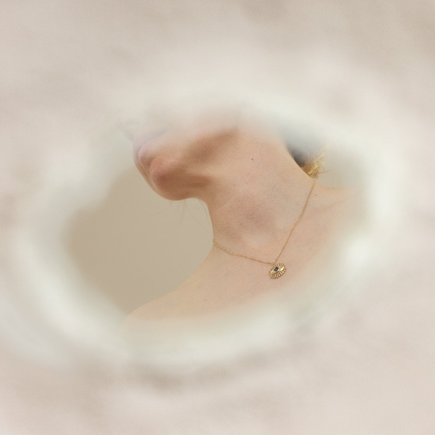 A delicate necklace with a gold pendant in an eye design set with a sapphire is worn by a model, shown through a soft, dreamy circular frame against a warm beige background.