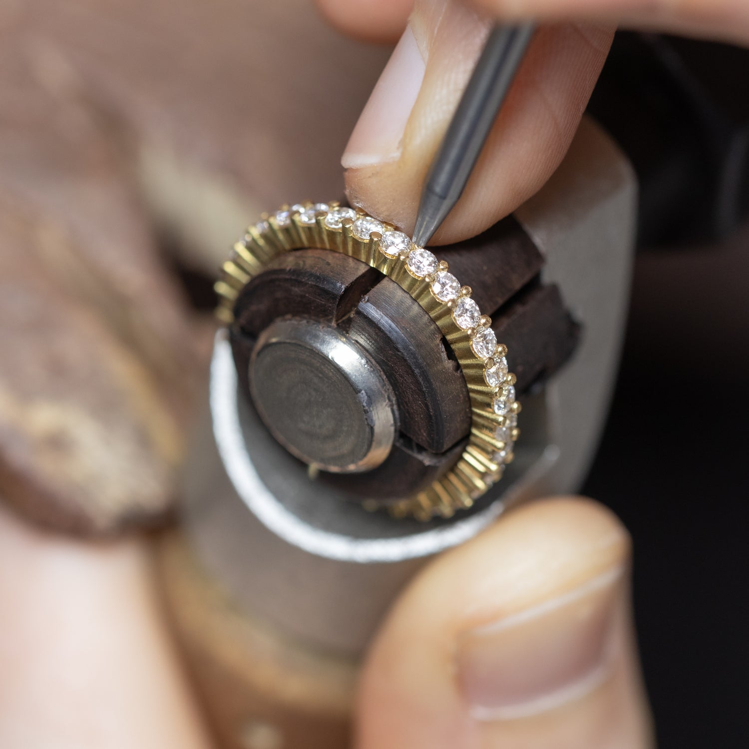 Close-up of a jeweller's hands using a precision tool on a gold eternity ring set with small lab-grown diamonds, held in a jeweller's setting tool.