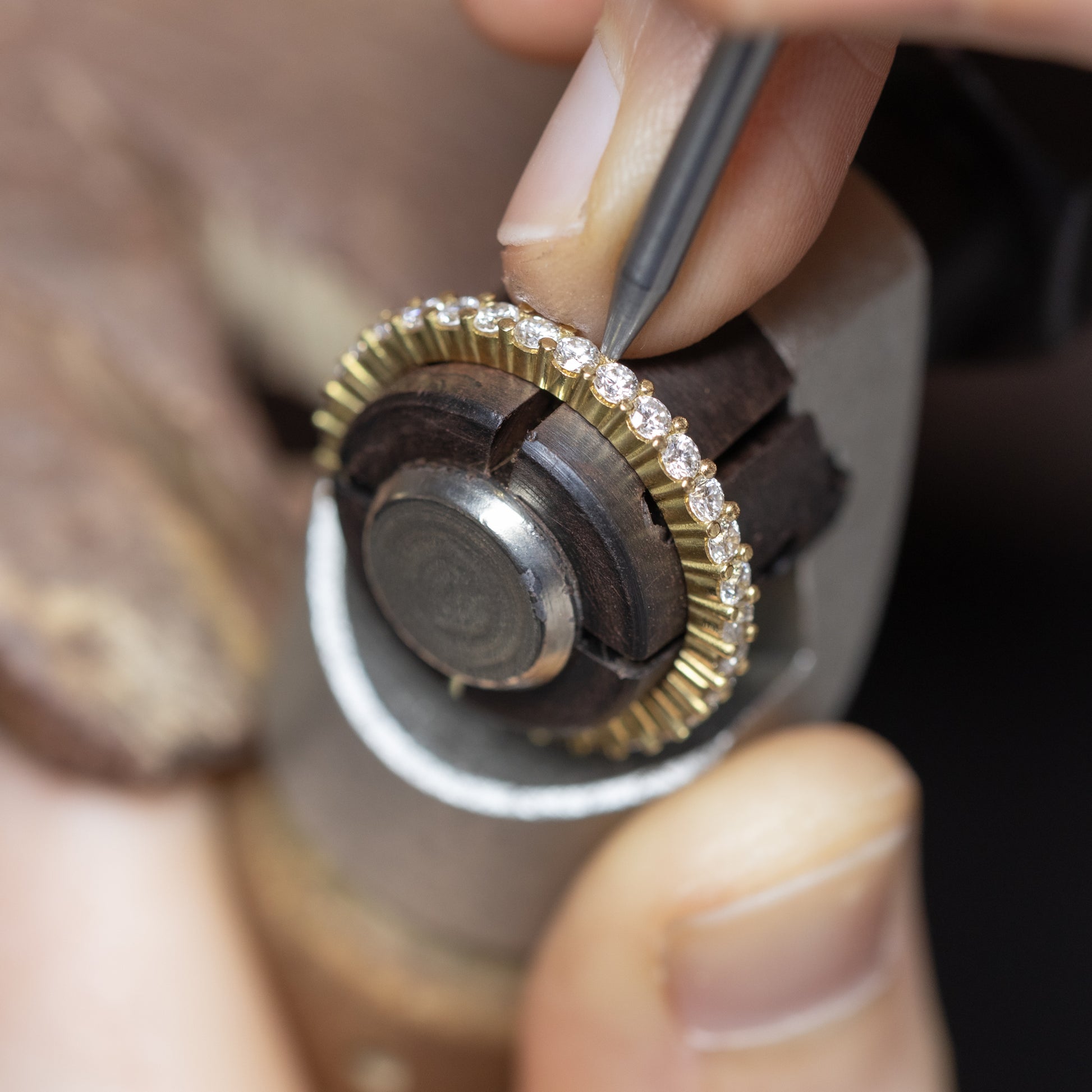 Close-up of a jeweller's hands using a precision tool on a gold eternity ring set with small lab-grown diamonds, held in a jeweller's setting tool.