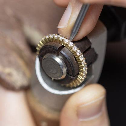 Close-up of a jeweller's hands using a precision tool on a gold eternity ring set with small lab-grown diamonds, held in a jeweller's setting tool.