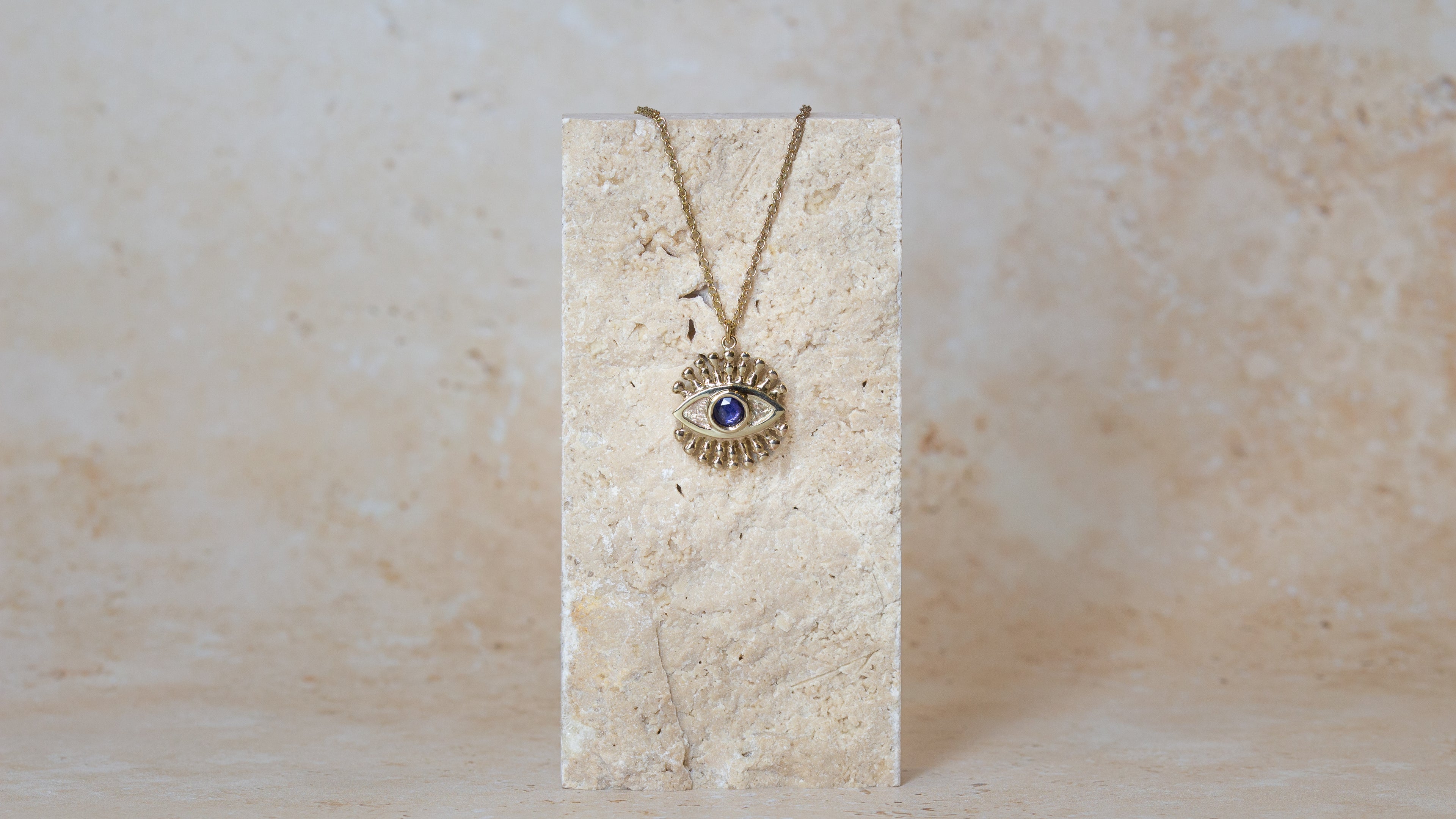 A gold eye pendant necklace set with a blue sapphire, hanging from a matching chain on a sandstone display block against a textured beige background.