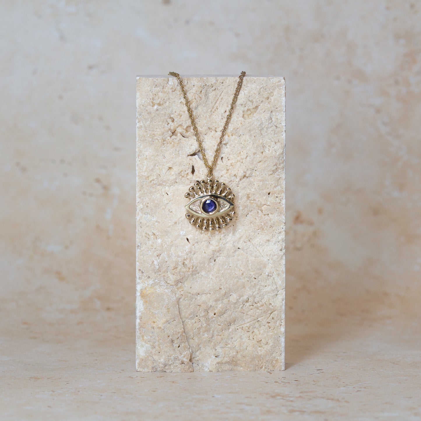 A gold eye pendant necklace with a sapphire iris, displayed on a textured sandstone block against a matching neutral background.