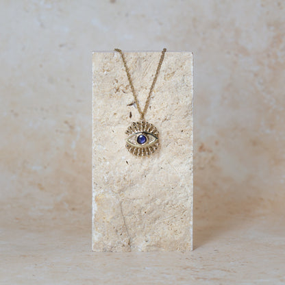 A gold eye pendant necklace with a sapphire iris, displayed on a textured sandstone block against a matching neutral background.
