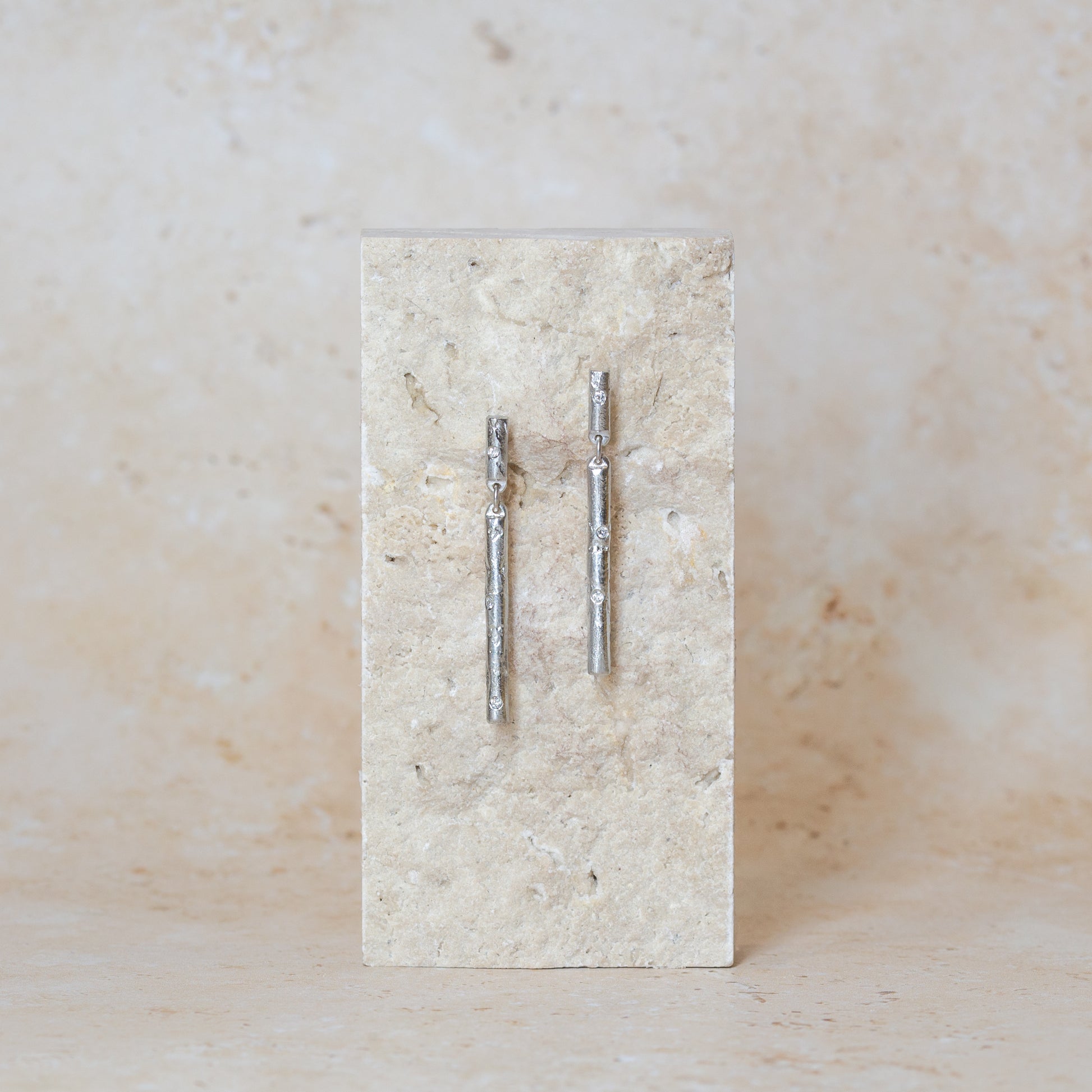Two silver bar stud earrings set with scattered lab grown diamonds displayed on a rectangular sandstone block against a matching textured background.