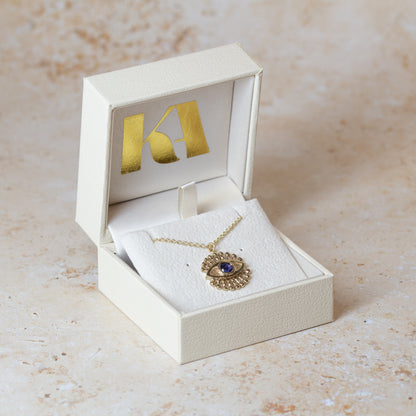 A gold eye pendant necklace with a blue sapphire, displayed inside an open cream-colored jewelry box with a gold foil Kate Archer logo on the inside lid, set against a light beige stone background.