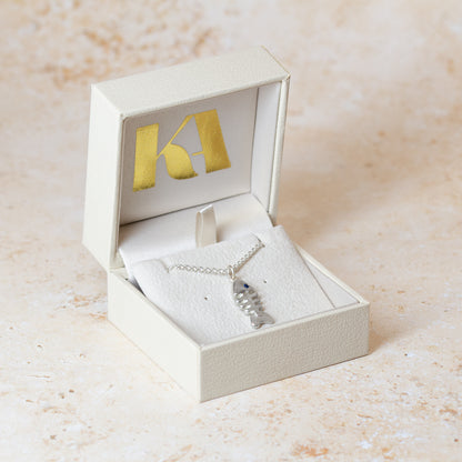 A silver fish-shaped pendant necklace with a sapphire set as an eye on a delicate chain, presented in an open coloured jewellery box with Kate Archer branding, set against a light beige textured background.