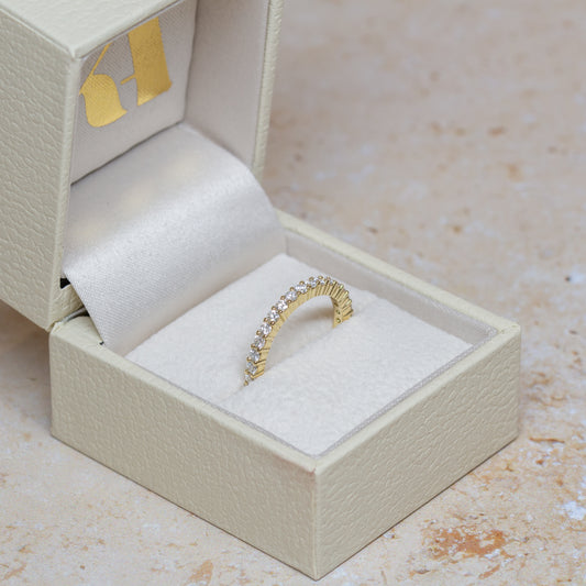 An 18cy yellow gold eternity ring set with 2mm lab-grown diamonds, displayed inside an open cream-colored textured jewelry box on a light beige stone surface.