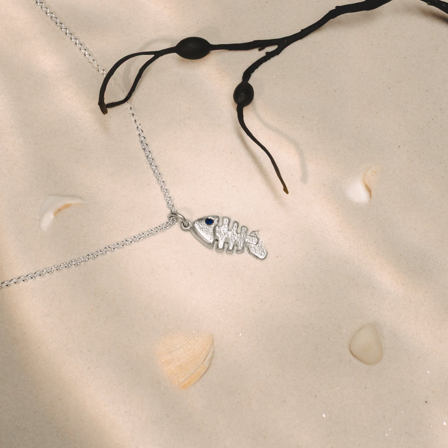 Silver necklace with a fish and sapphire pendant on a sand surface with seashells.